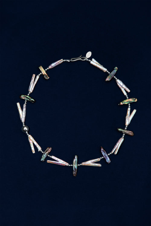 A circular beaded necklace featuring iridescent, white and black irregular pearls, silver beads, displayed against a dark navy blue background.