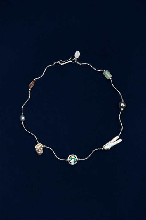A delicate sterling silver wave necklace with multiple small pearl and stone accents in various colors, including a central abalone shell displayed on a dark navy blue background.