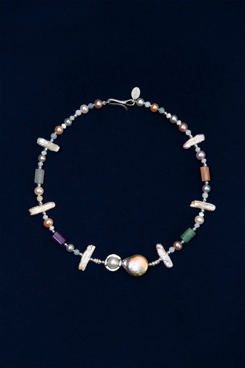 A beaded necklace featuring a large iridescent pearl, silver-toned beads, small multicolored pearls, white stick pearls, and semi-precious stones displayed on a dark navy background.